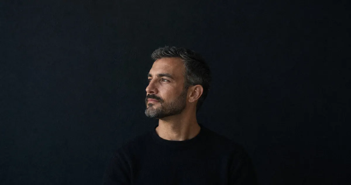 Retrato de tres cuartos de un hombre de unos 45 años con barba corta canosa y línea de barba nítida, sobre fondo oscuro