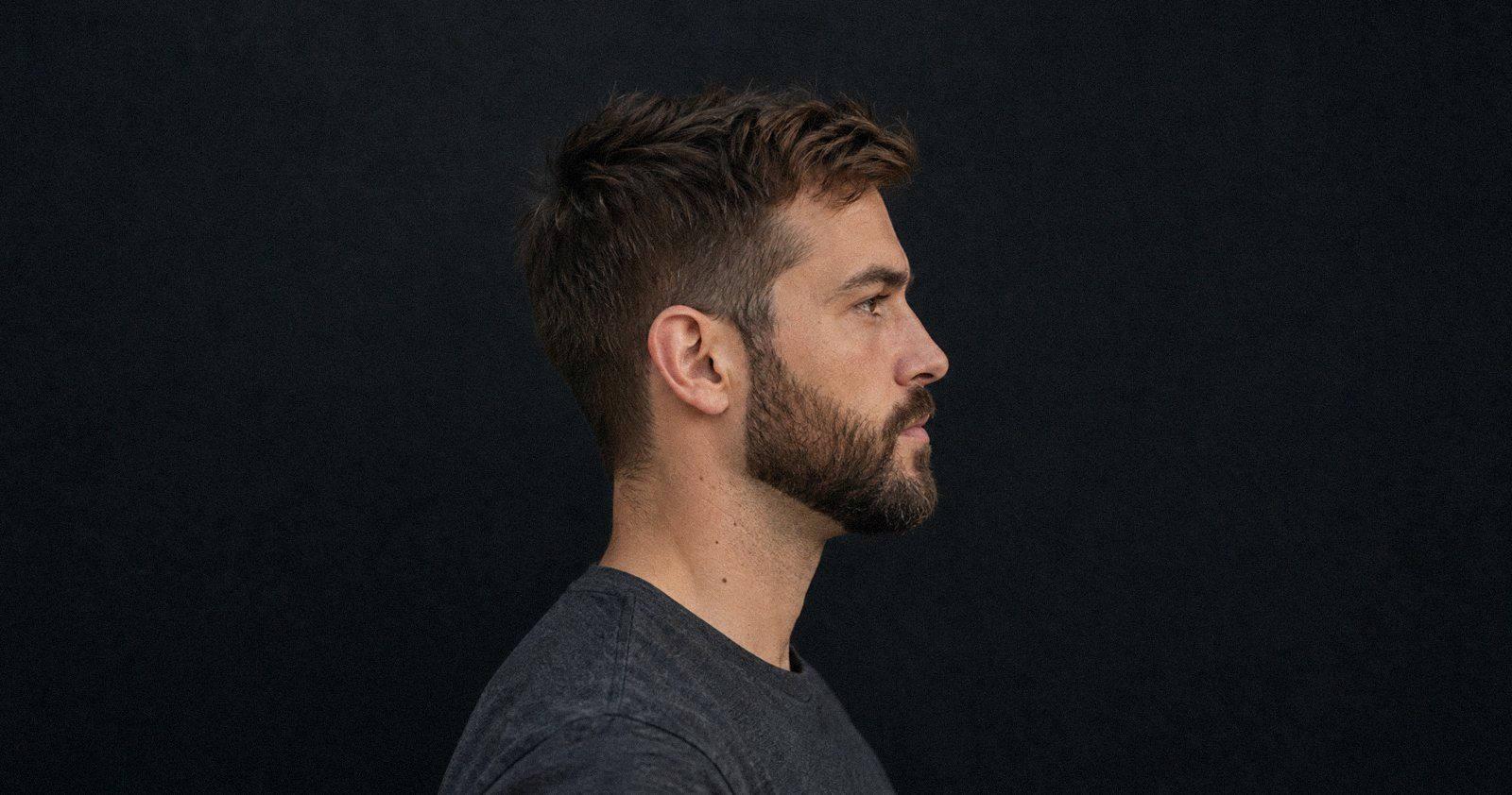 Perfil de un hombre de unos 35 años con barba poblada perfilada que marca el ángulo mandibular, camiseta antracita