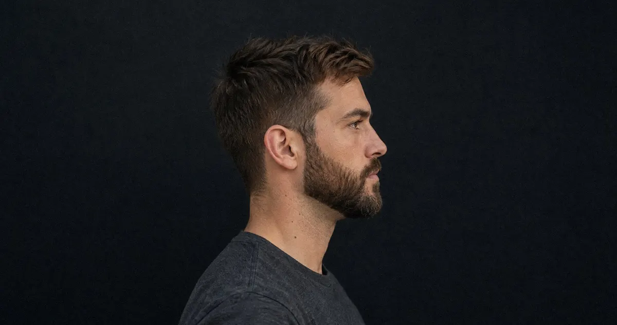 Perfil de un hombre de unos 35 años con barba poblada perfilada que marca el ángulo mandibular, camiseta antracita